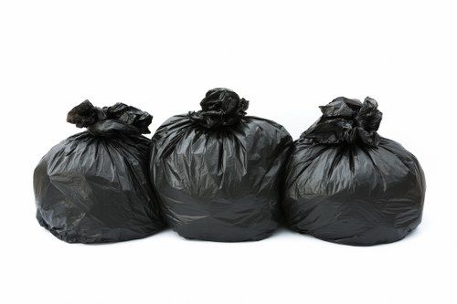 Three black rubbish bags made of thick, shiny plastic material are arranged in a row against a plain white background. The bags are slightly crumpled and tied at the top, with visible wrinkles and creases on their surface. They appear filled and are of similar size, suggesting they contain household waste or debris ready for disposal. The plain background emphasizes the bags without any additional context, aligning with rubbish removal services offered by East Ham House Clearance. The image focuses solely on the waste bags, likely in preparation for collection or transportation, typical of domestic or small-scale clearance operations within the East Ham area or nearby postcode regions. The lighting is even and neutral, providing clear visibility of the bags' texture and color, suitable for illustrating rubbish collection services on a professional website.