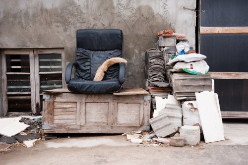 A black executive office chair with cushioned seat and backrest, featuring armrests, is positioned on top of a weathered wooden chest or storage box placed on the ground. To the right of the chair, there is a pile of construction debris and waste materials, including grey concrete blocks, white broken tiles or panels, and various discarded packaging and fabric coverings. The setting appears to be an outdoor area or alleyway with a concrete surface, adjacent to a rough, weathered wall with visible stains and graffiti marks. Ventilation grilles are partially visible near the ground behind the debris, and a black metal door or shutter is seen on the far right. This scene is likely associated with waste clearance or rubbish removal services in the East Ham area, provided by East Ham House Clearance, highlighting common clutter found during clearance or renovation projects.