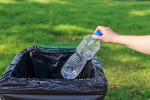 A person's hand is seen placing a clear plastic water bottle into a black rubbish bin lined with a black plastic liner, situated outdoors on a grassy area. The bottle is partially squeezed, with the person's arm extending from the right side of the image, and the background shows vibrant green lawn with a slightly blurred natural setting. The scene reflects typical waste disposal activity, relevant to rubbish removal services provided by East Ham House Clearance, in a residential or community outdoor environment. The image emphasizes the action of proper waste segregation and collection, highlighting the importance of recycling and rubbish disposal in urban areas like East Ham, London.